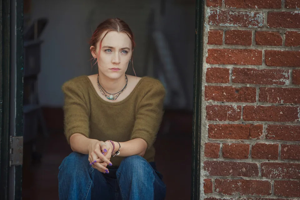 Person sitting pensively at a doorway, wearing a sweater and jeans, with their hair tied back. Brick wall visible in the background