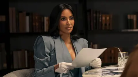Disney+ Picture of Kim Kardashian starring in All's Fair on Disney. She is wearing a grey suit jacket with huge shoulder pads and white gloves.