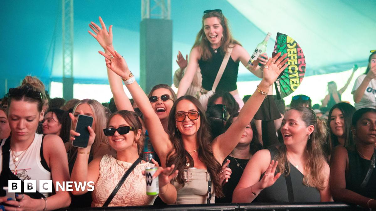 A crowd of people in a big festival tent with hands up and smiling into the camera. Someone is up on their friends shoulders and one girl is holding a "defected records" fan.