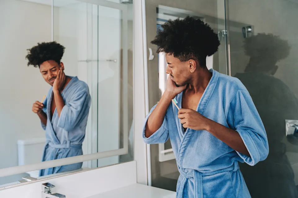 Person in a bathroom wearing a robe, brushing their teeth while looking in the mirror