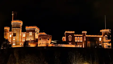 Duncan Walker A grand stone castle is dramatically illuminated in warm golden lights against a pitch-black night sky. Its towers and battlements stand out with sharp architectural details, creating a striking silhouette.