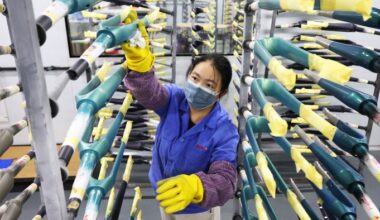 A worker wearing a mask and gloves inspects carbon fibre bicycle frames arranged on racks in a factory setting.