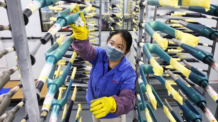 A worker wearing a mask and gloves inspects carbon fibre bicycle frames arranged on racks in a factory setting.