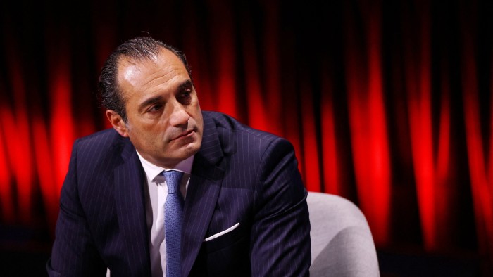 Antonio Filosa sits in a chair wearing a dark pinstripe suit and light blue tie, with a red curtain in the background.