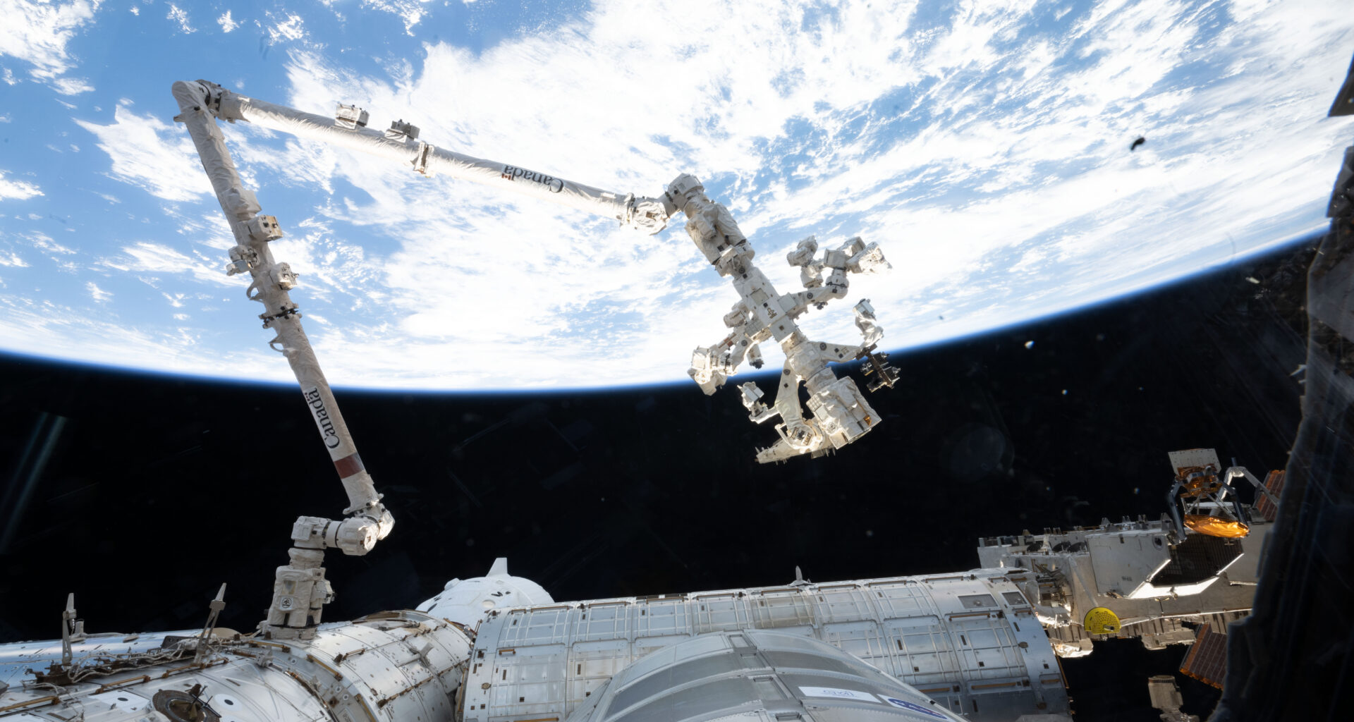 The 57.7-foot-long Canadarm2 robotic arm extends from the International Space Station. The arm is long, cylindrical, and white, with several joints that are bent to the right (at left in the image) and downward (at center). Earth makes a bright white and blue background for the top half of the photo. Our home planet contrasts starkly with the darkness of space.