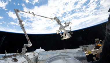 The 57.7-foot-long Canadarm2 robotic arm extends from the International Space Station. The arm is long, cylindrical, and white, with several joints that are bent to the right (at left in the image) and downward (at center). Earth makes a bright white and blue background for the top half of the photo. Our home planet contrasts starkly with the darkness of space.