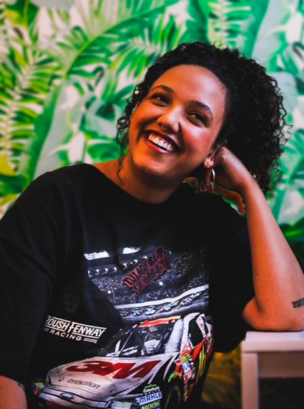 A young woman with curly black hair in a high ponytail and a black t-shirt laughs off to the side. She is resting her face on her hand at a white table, in front of leafy green wallpaper.