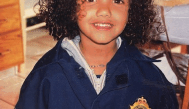 A smiling little girl with chin-length curly hair wears an oversized dark blue jacket with a badge on the front.  .