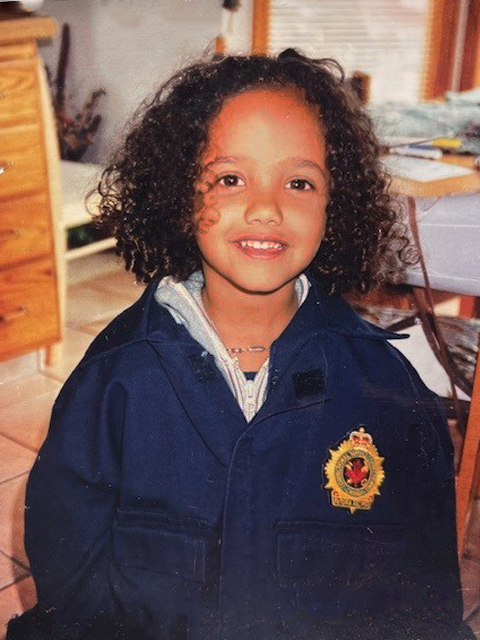 A smiling little girl with chin-length curly hair wears an oversized dark blue jacket with a badge on the front.  .