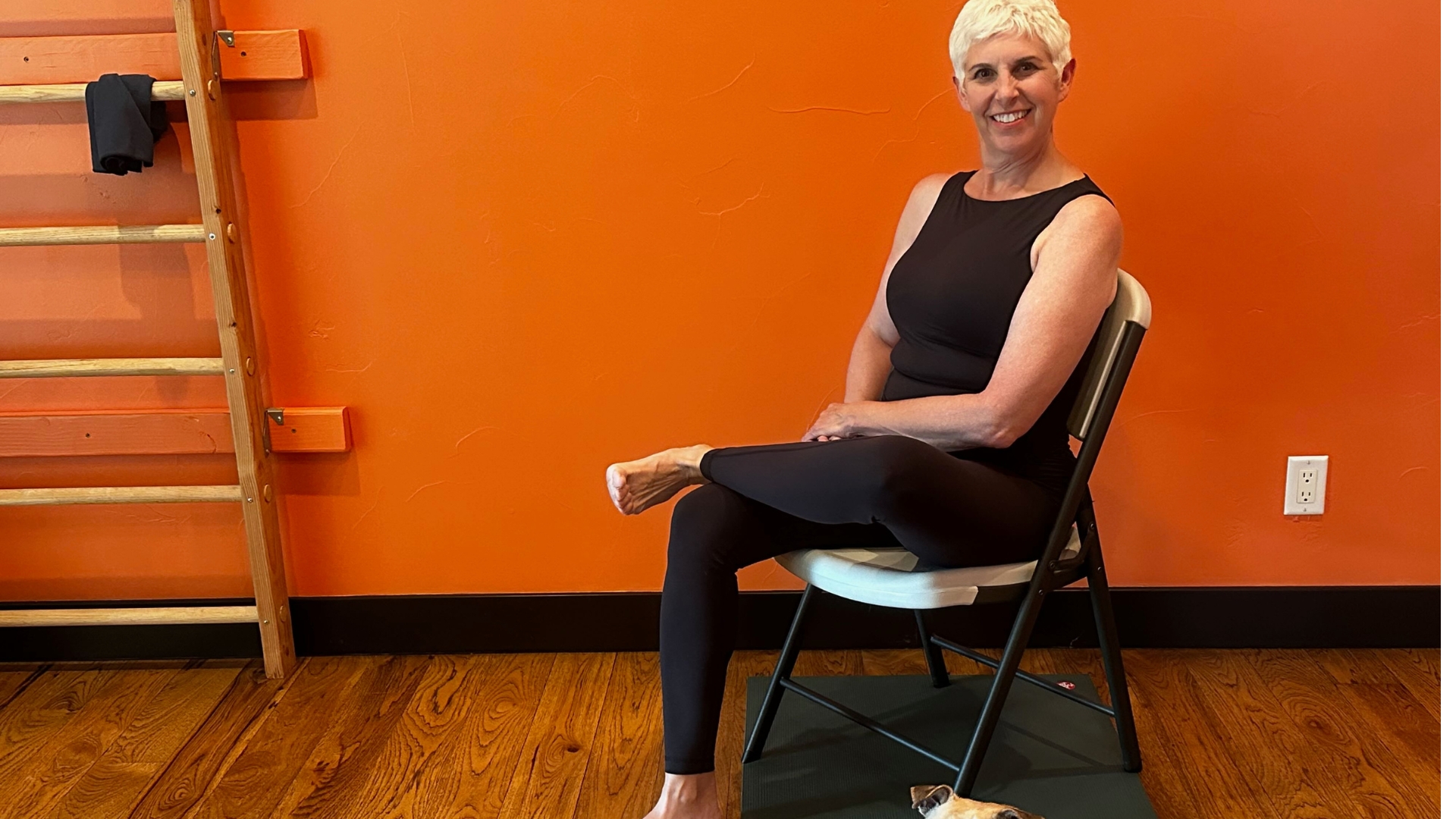 woman on a chair with one ankle over her other thigh. there's an orange wall behind her.
