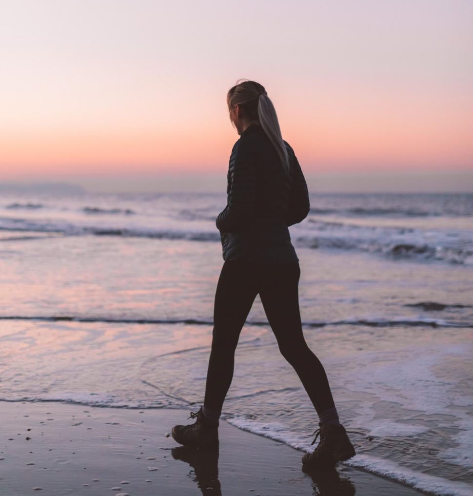 Person in a jacket and leggings walks along the shore at sunset, staying just one meter from the waves, with a colorful sky in the background.