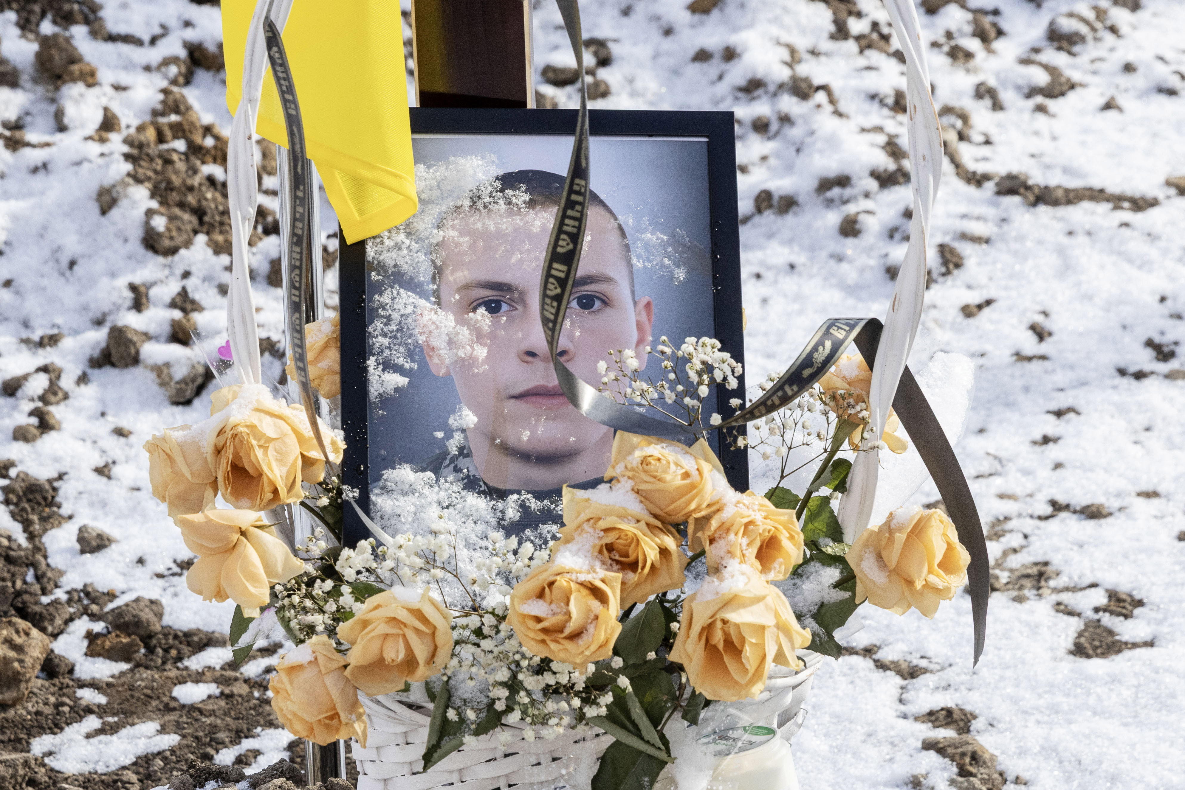 A framed photograph of a young man with snow on its surface, yellow roses, and a yellow ribbon, at a grave in Lviv cemetery.