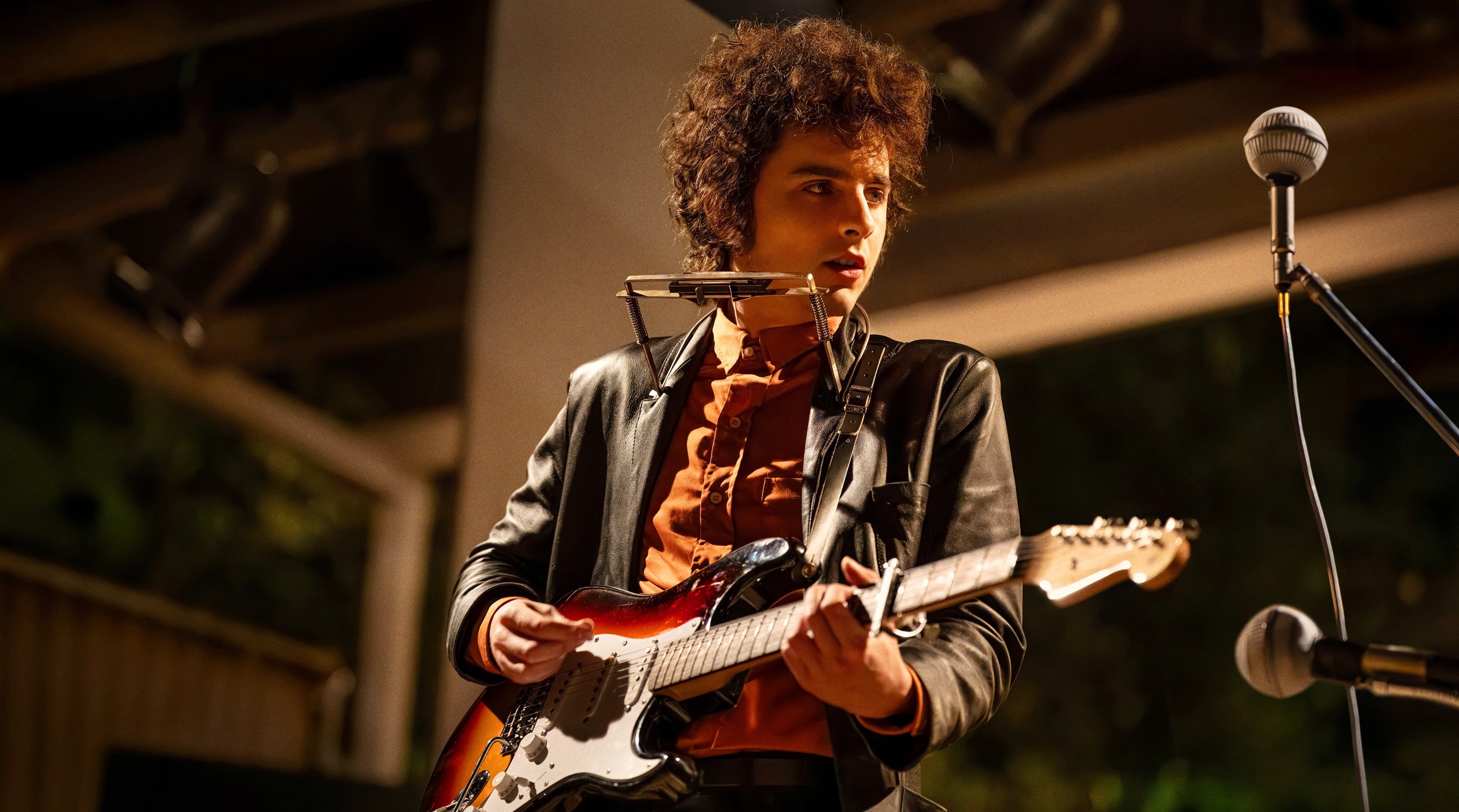 Timothee Chalamet as Bob Dylan in A Complete Unknown, playing guitar and harmonica.