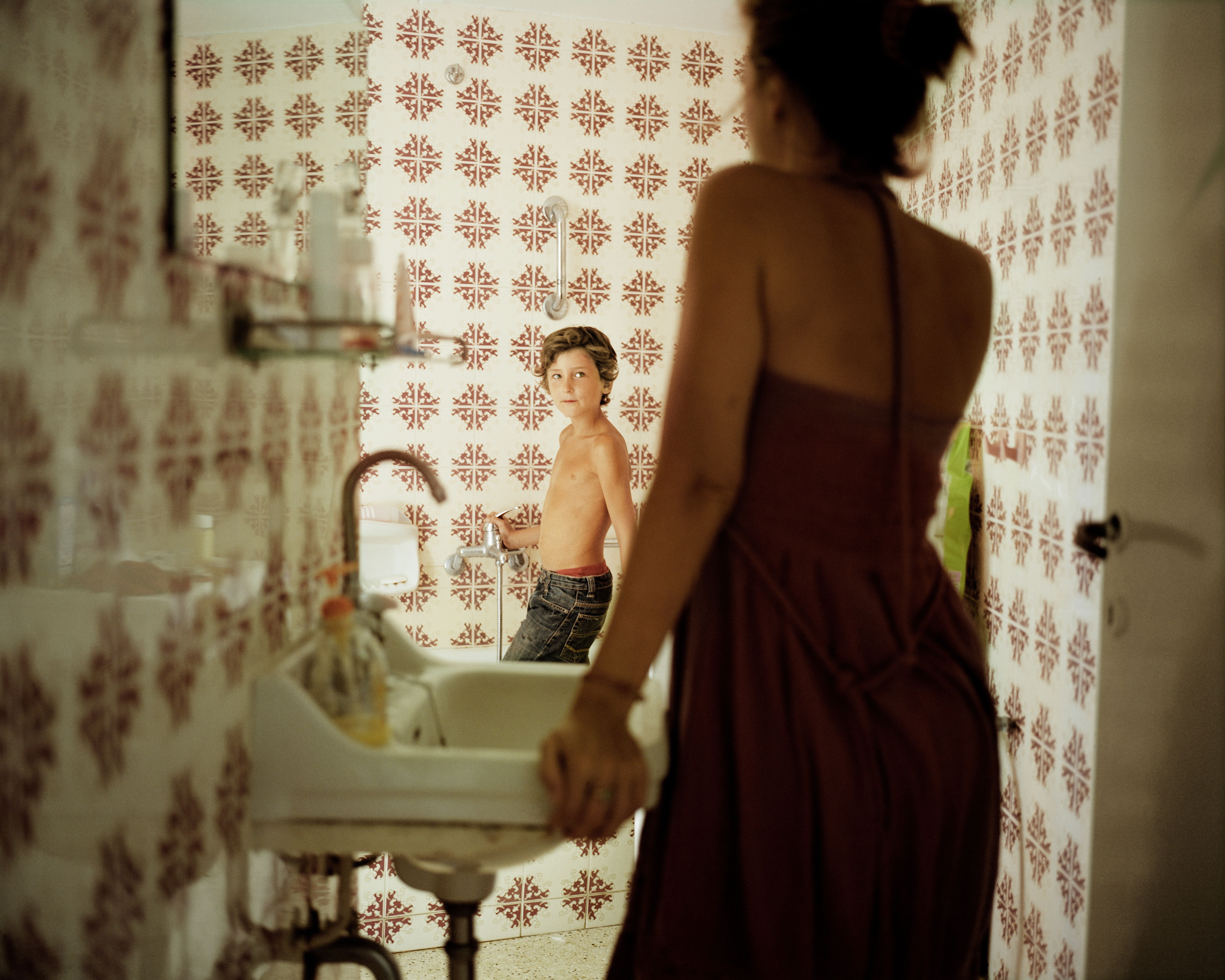 A woman in a dark dress stands with her back to the camera, looking into a mirror. Her son, shirtless and in jeans, looks back from the mirror.