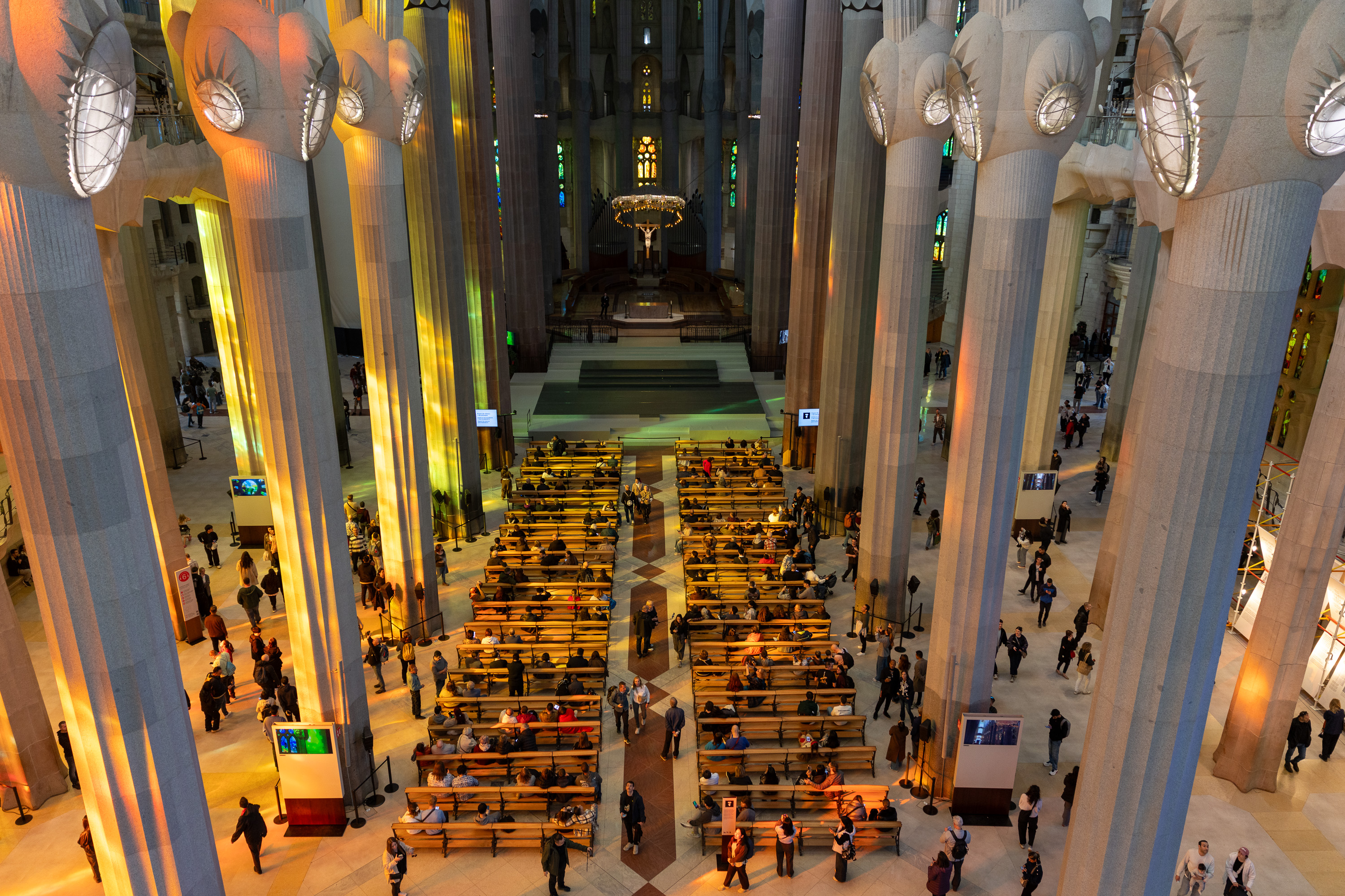 Interior view of the Sagrada Familia basilica with visitors, rows of wooden pews, and massive columns reflecting colorful light.