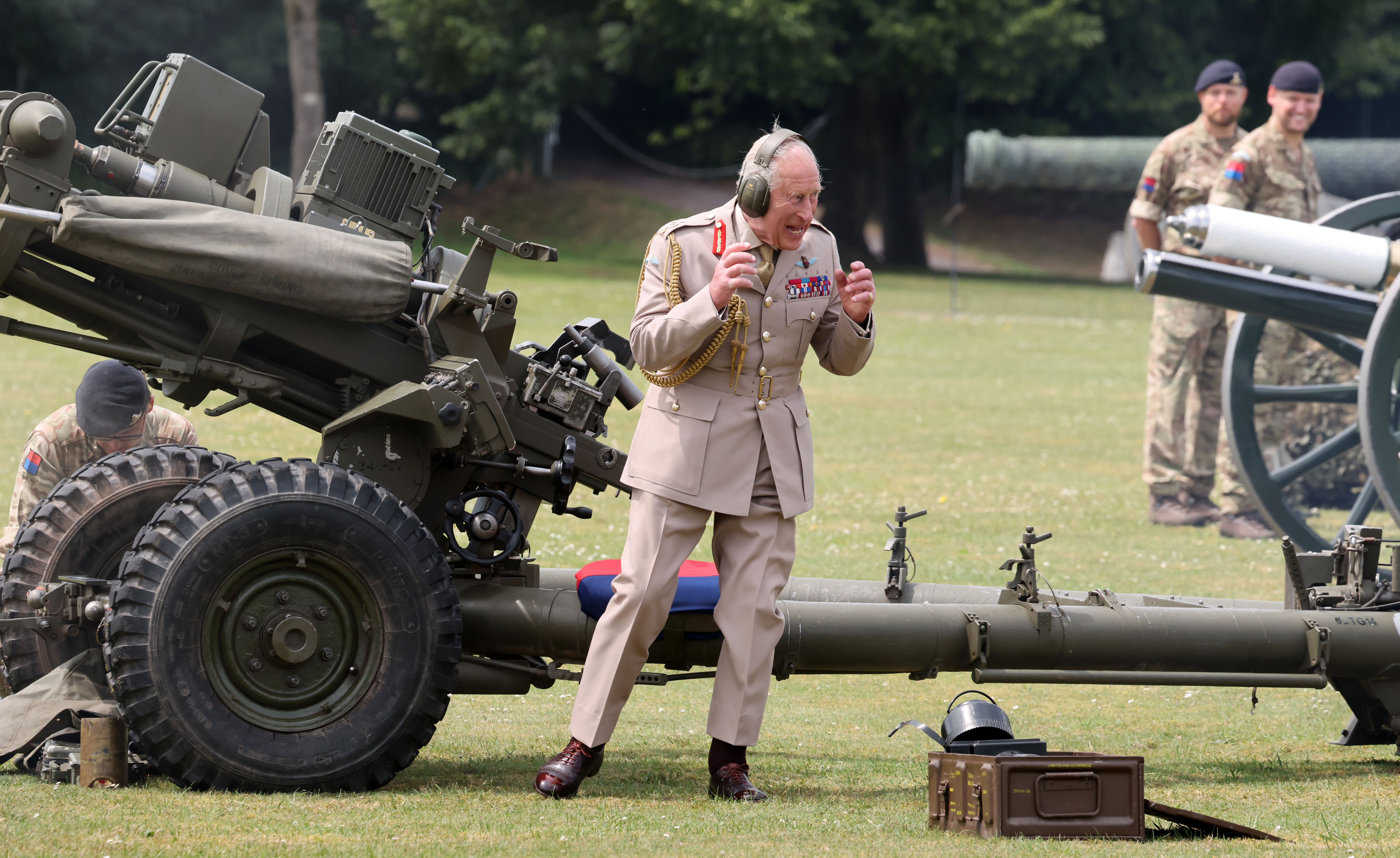 King Charles III in military uniform and earmuffs, reacting to firing a 105mm artillery piece.