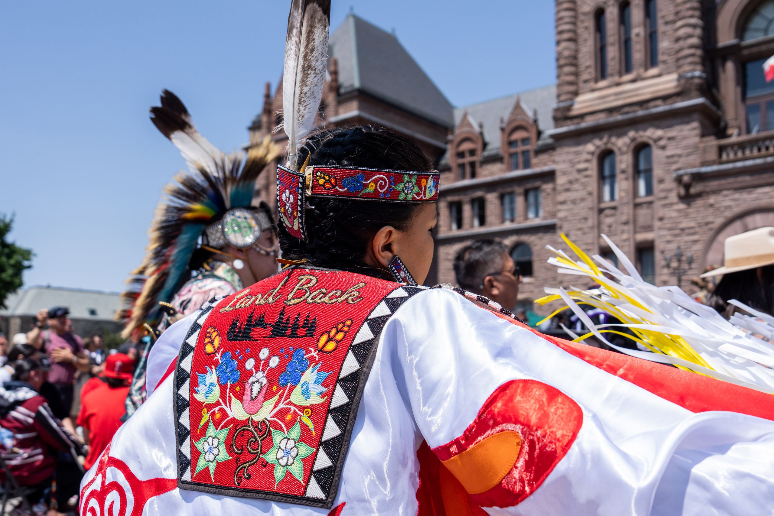 Person in traditional garb seen from the back with the phrase "land back" embroidered on their shirt