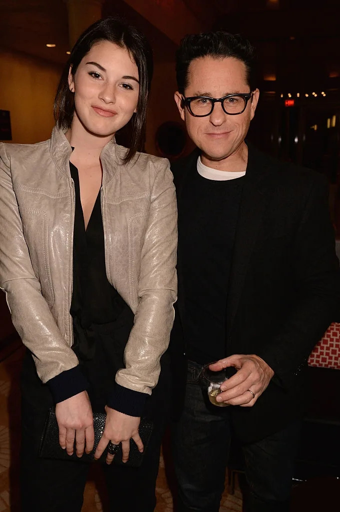 Two people posing together at an event; one wears a casual jacket over a dress, the other in a simple sweater and glasses, holding a drink