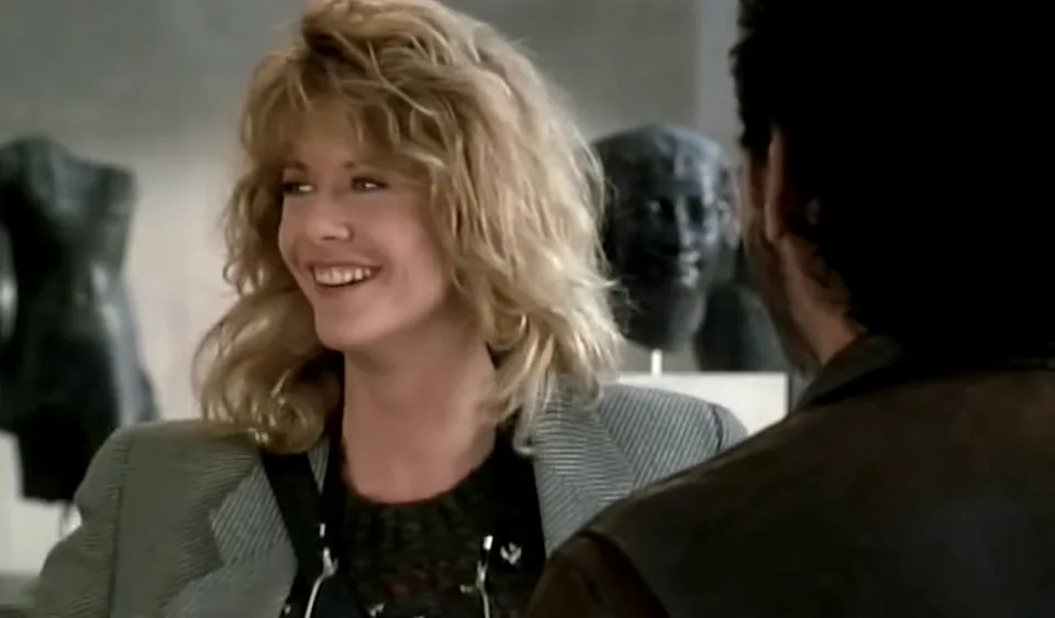 Woman with wavy hair smiles while talking to a man, wearing a textured top and tailored blazer. Background features abstract sculptures