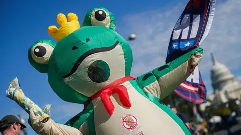 Getty Images A protester in a frog costume holds a subway sandwich and wears a sticker that says No Kings