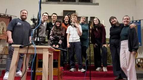 The Inner Voices A group of people are standing shoulder to shoulder inside a large church, with recording equipment in front of them.