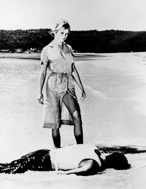 Marka/Universal Images Group via Getty Images Brigitte Bardot in a still from And God Created Woman, wearing a dress that is partly unbuttoned from the hem so it parts to reveal her thigh, as she stands over a man lying face down on a beach.