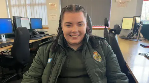 BBC A young woman with dark hair in a pony tail is sat in a chair in the emergency call centre. She is wearing a dark green ambulance jacket and t-shirt, and is smiling to the camera. 