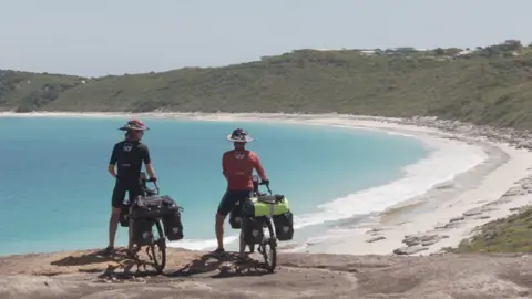 Kohler family Two men holding bikes face away from the camera as they overlook a sweeping sandy bay and a turquoise sea. 