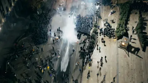 Shutterstock Crowds scatter as water is sprayed out from trucks in the Georgian capital - there is a large plume of water and widely dispersed mist. It is dark and the shot is taken from above.