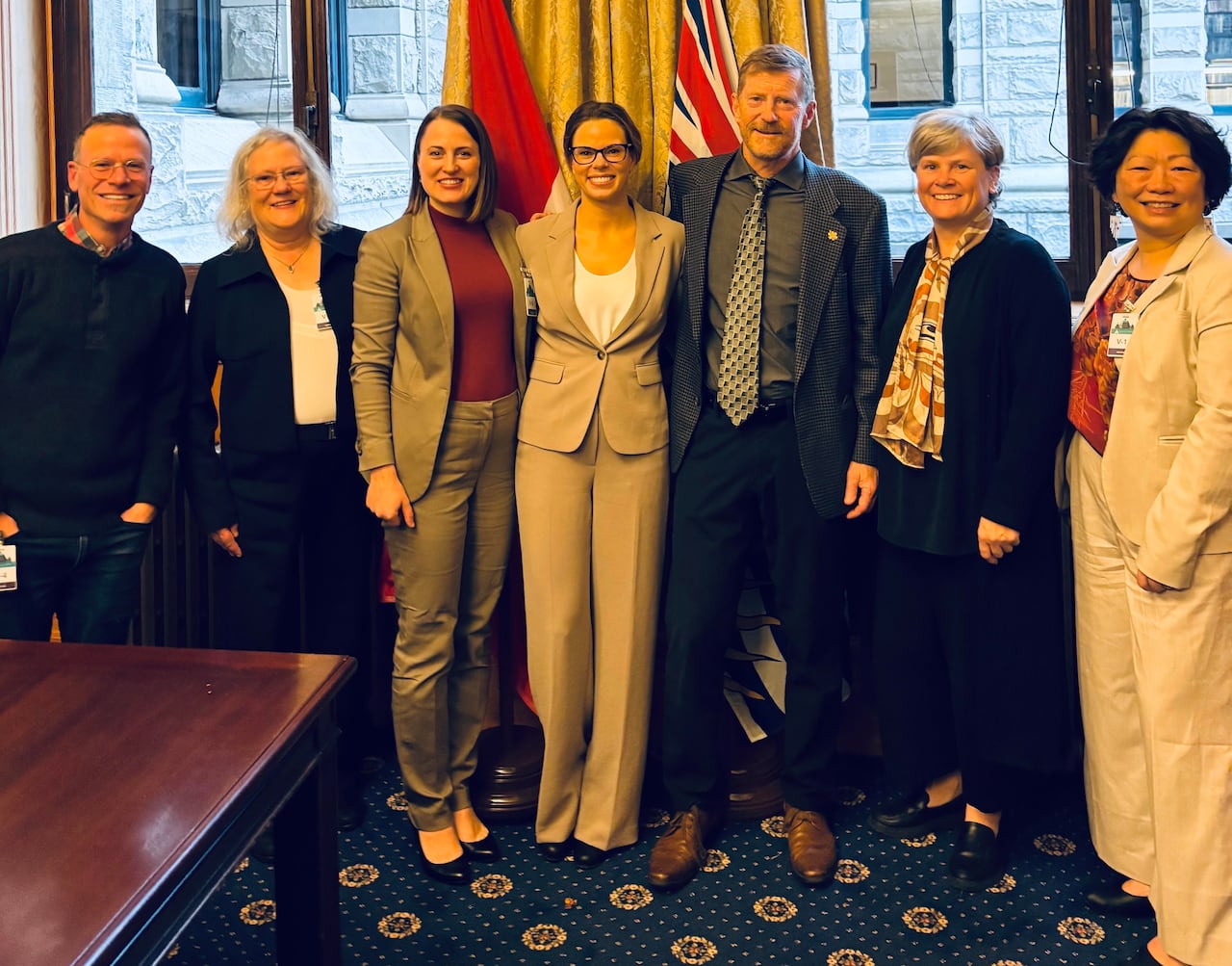 A group of seven people in formal clothes stand with arms across eachother's backs to pose for a photograph in front of a wood-framed window.