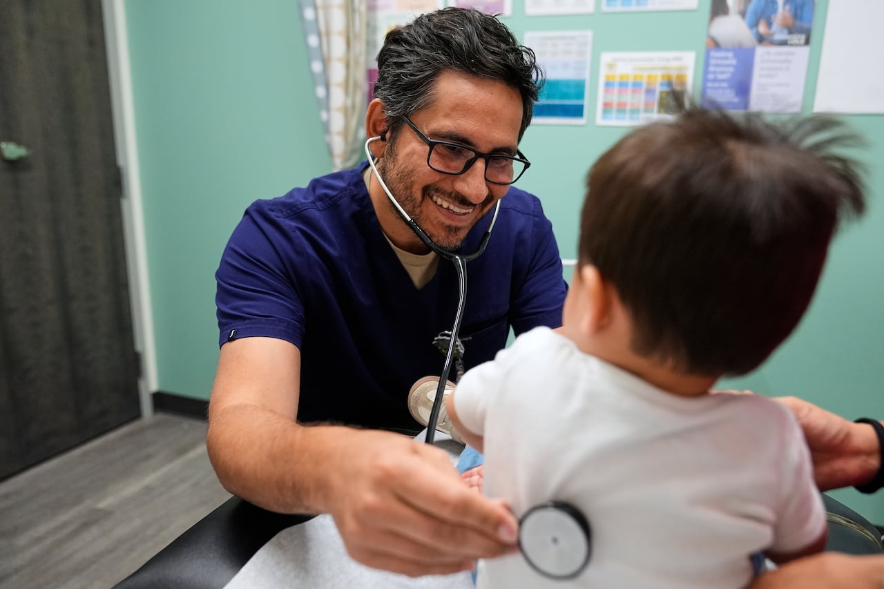 A pediatrician smiles as he listens through a stethoscope held against the back of a 16-month-old boy. 