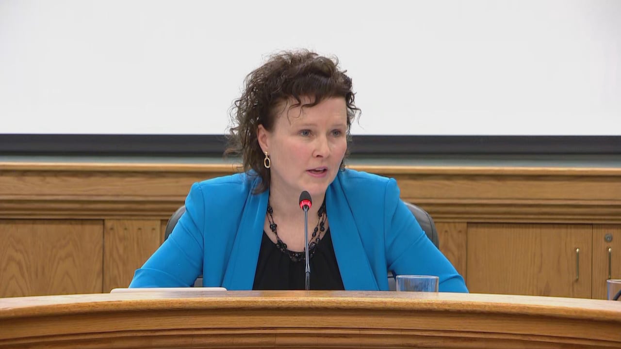 A woman in a blue suit sits at a wooden desk.
