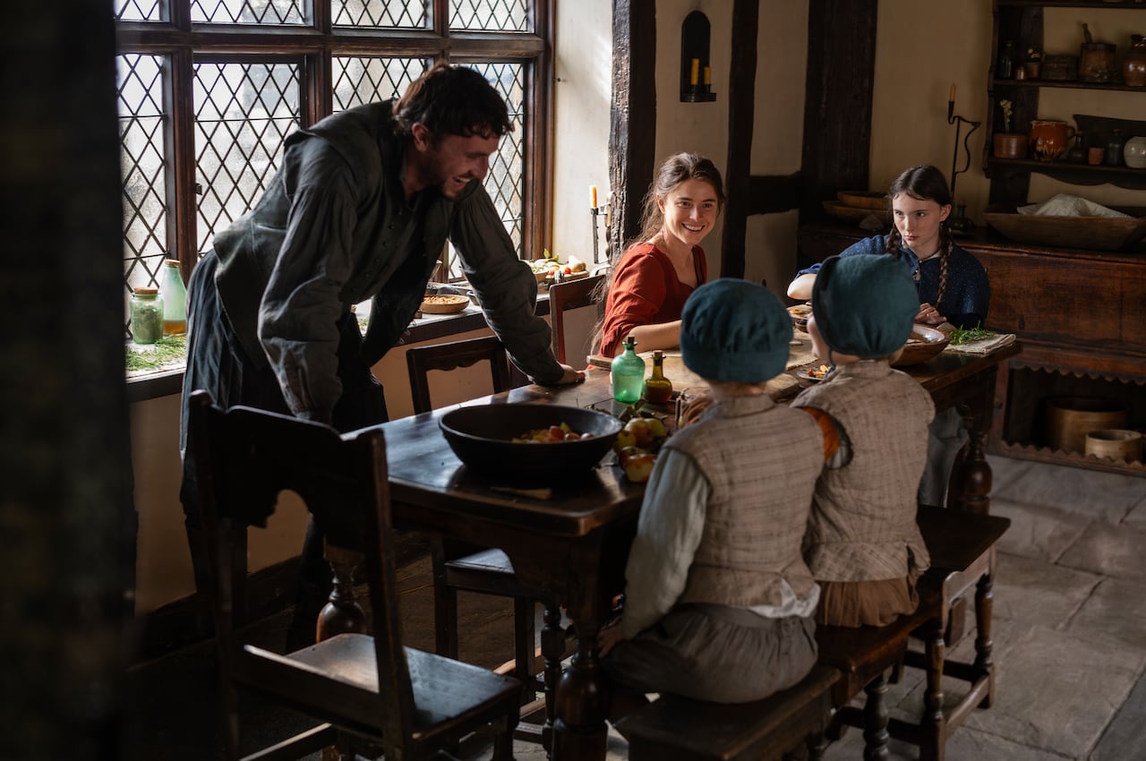 A smiling man in period-dress leans over a table. Also sat at the table is a smiling woman, young teenage girl and two children. 