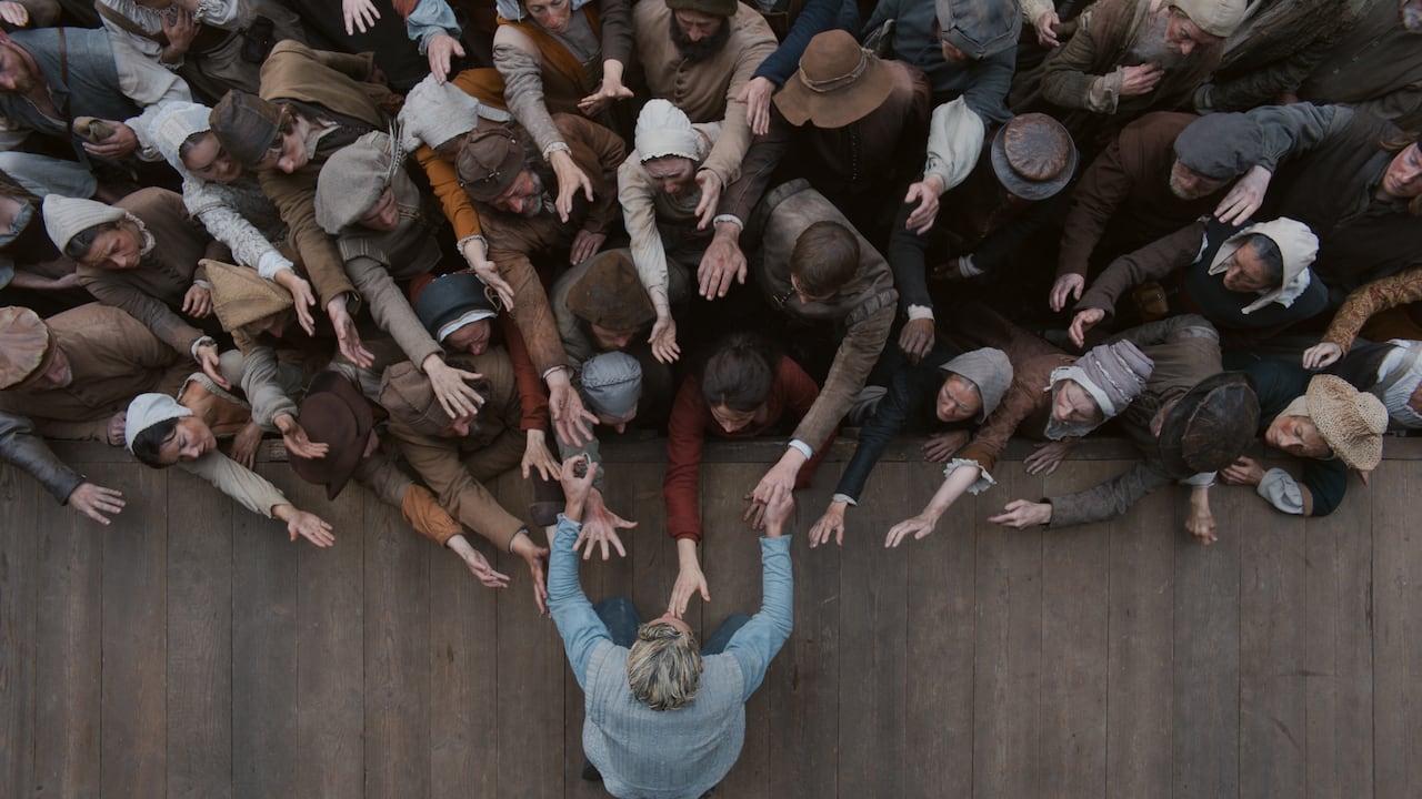 A top-down shot shows a man on a stage, leaning over toward an audience. Both the man on the stage, and the audience members have their hands outstretched to one another.