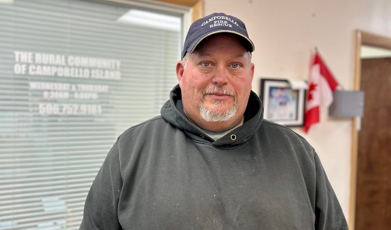 A man with a ball cap and a grey hooded sweater looks at the camera with a serious face, while standing inside an office area. 