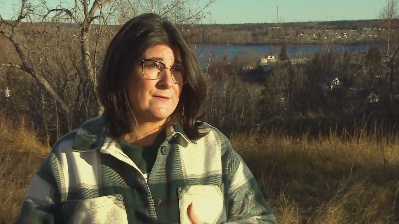 Woman with dark hair and eyeglasses wears a blue and white plaid jacket and stands beside a lake