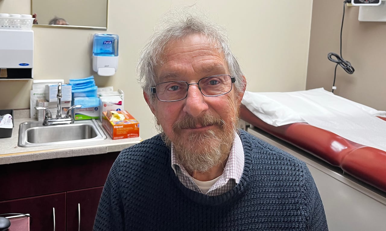 Man with white hair and white beard wears eyeglasses and a blue sweater while sitting in a doctor's office