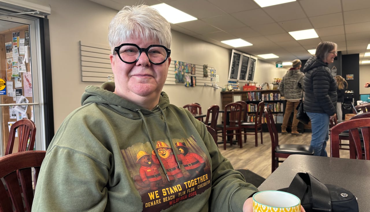 Woman with short white hair and bold black eyeglasses wears a green hooded sweatshirt in a coffee shop