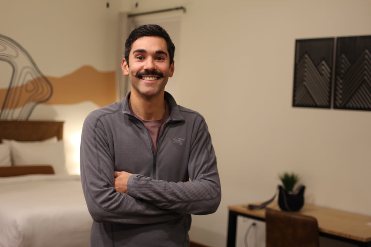 A man with a mustache in a zip-up sweater poses for a photo inside a hotel room.