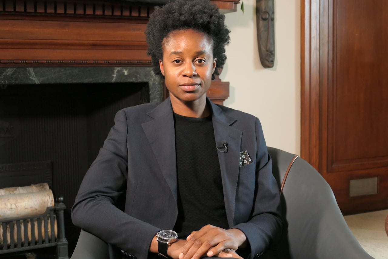 A person in a dark suit sits on a chair with their hands folded.