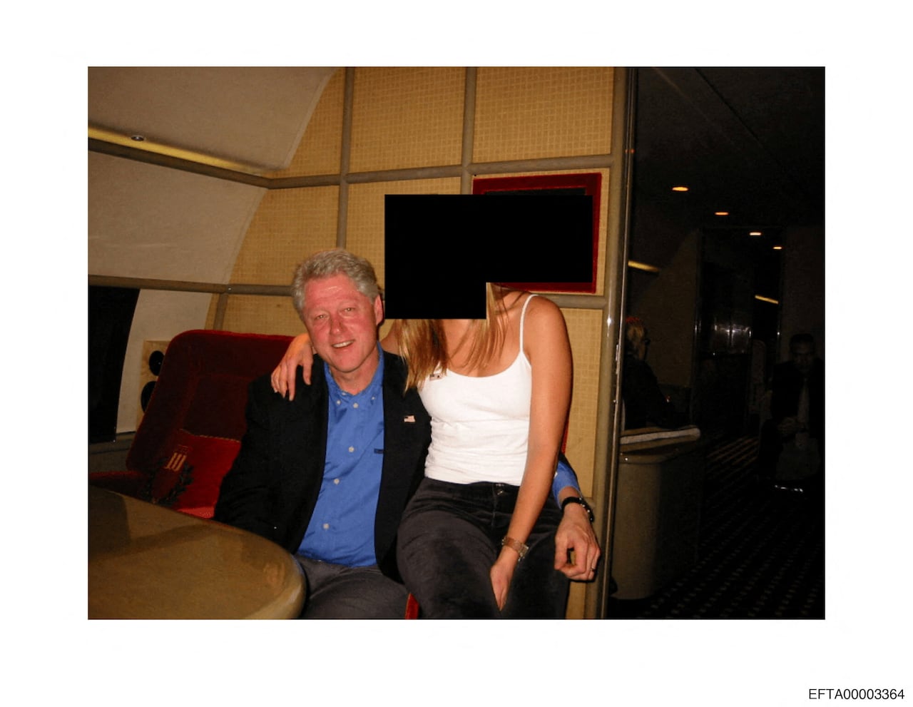 A grey-haired man is sitting in a seat on a private plane. A blonde woman in a white tank top sits on the arm of the chair with her arm around his shoulders. Her face is redacted.