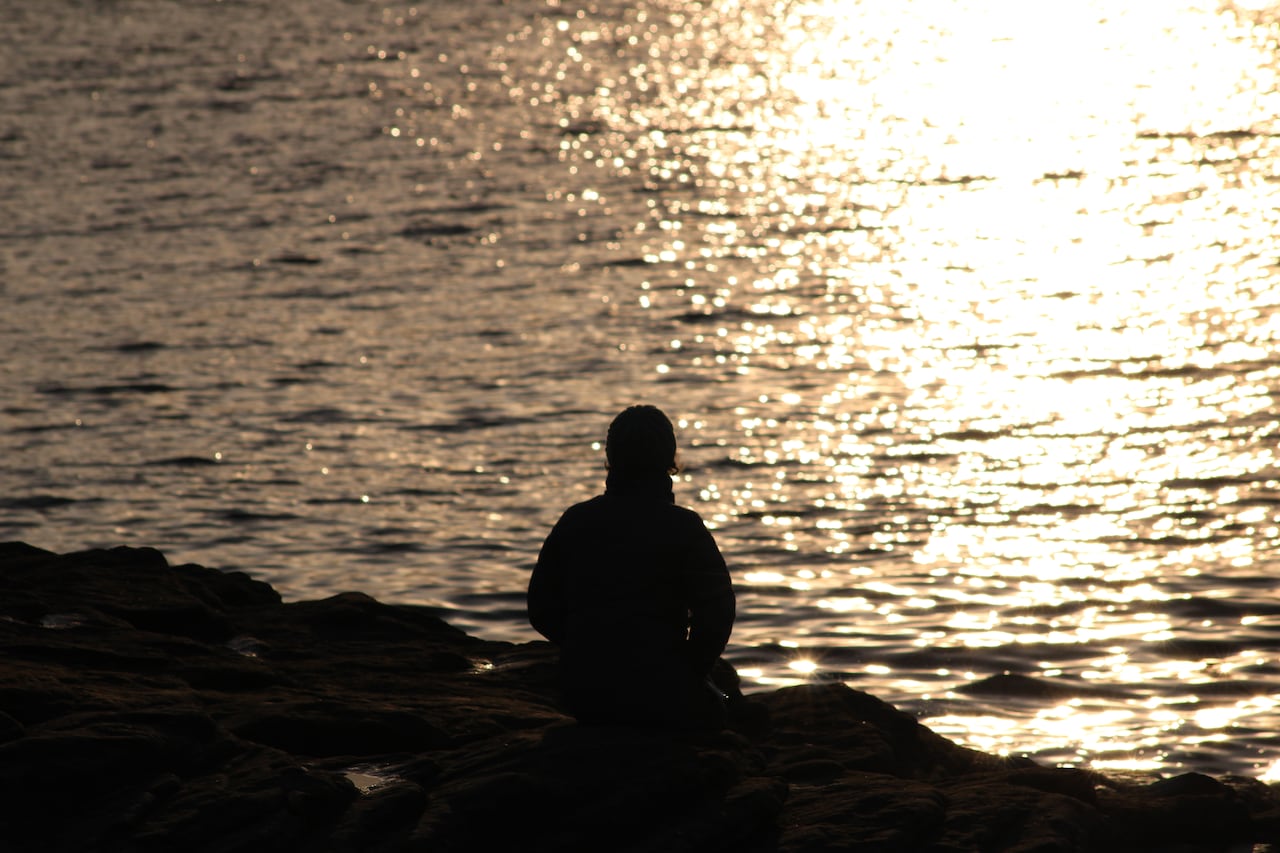 A woman sits on a rock on a beach looking at the ocean.