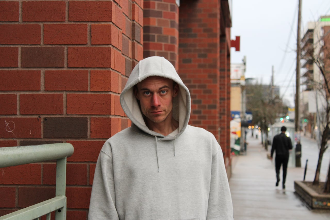 white man in grey sweatshirt with hood up stands against red brick wall and looks directly at camera