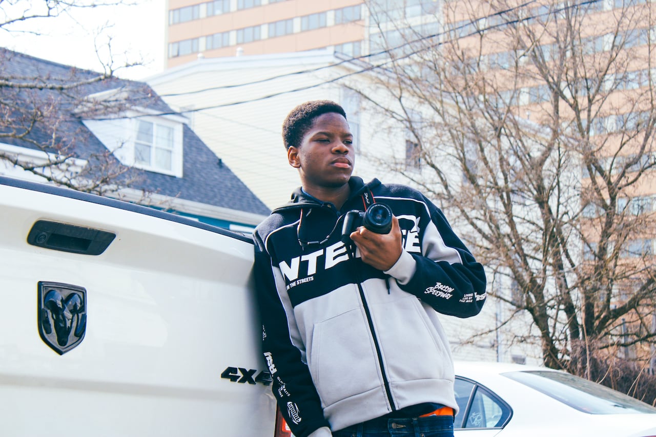 Black boy holding a camera leans against a white truck