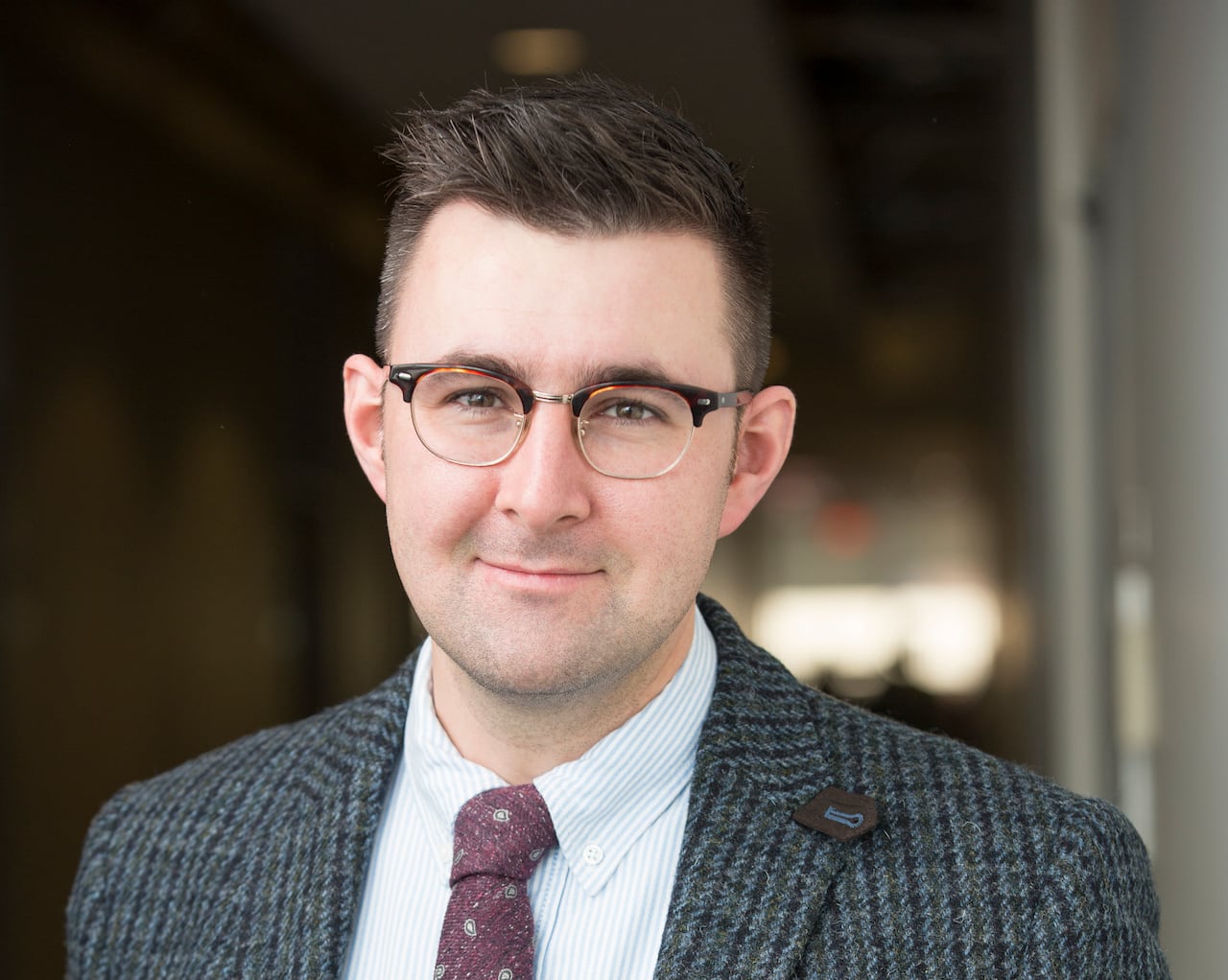 A man in a checkered blazer, maroon tie and glasses looks into a camera.