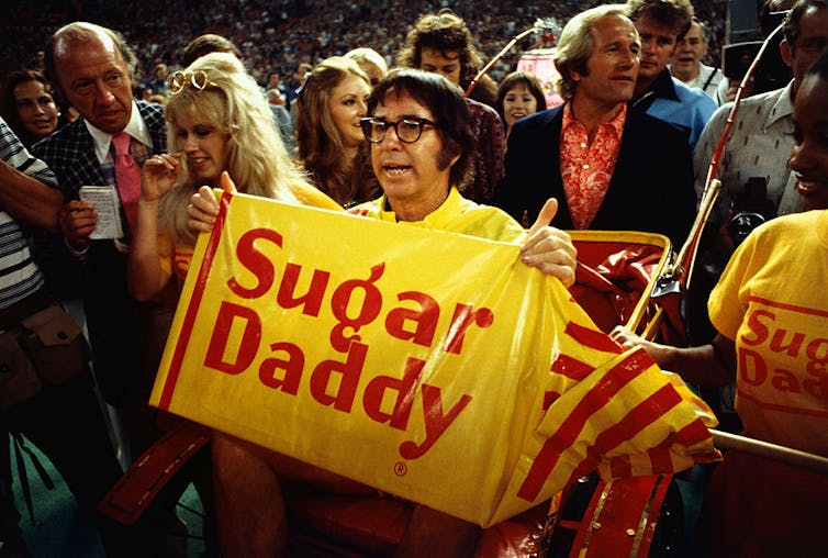 Man wearing glasses holds a yellow sign reading 'Sugar Daddy.'