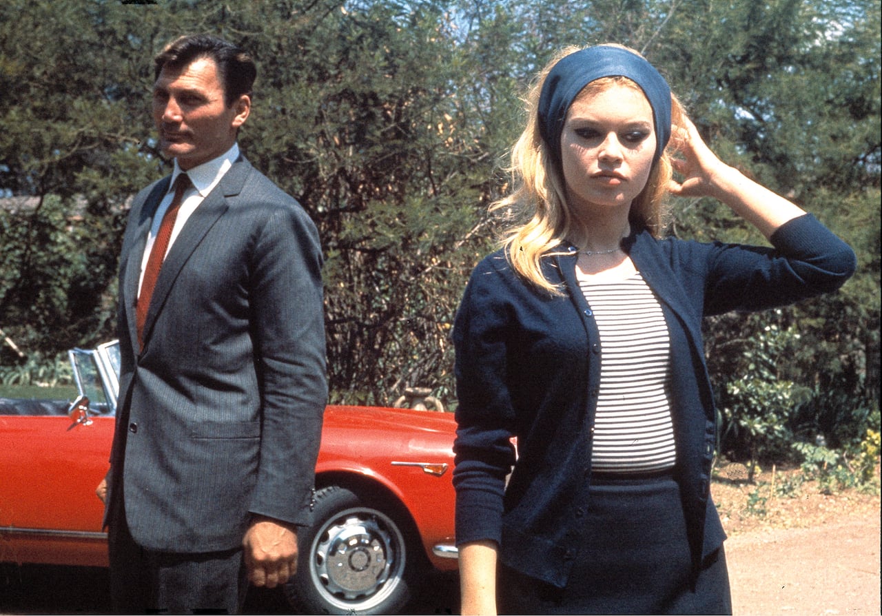 A woman and a man stand in front of a car in the 1960s.