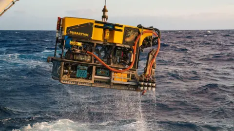 Schmidt Ocean Institute A large yellow submersible vehicle is lifted from the choppy ocean waters.