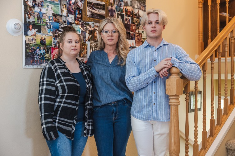 A woman stands in between two younger people. They are posing in front of a poster covered in family photos.