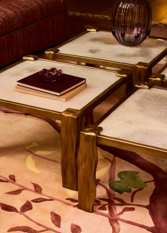 Three nested square coffee tables with gold frames and glass tops on a floral carpet, featuring stacked books and a vase.
